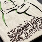 Крупним планом гравійований напис «Коханій Матусі. З любов’ю!» на стрічці з квітами і силует мами з малюком